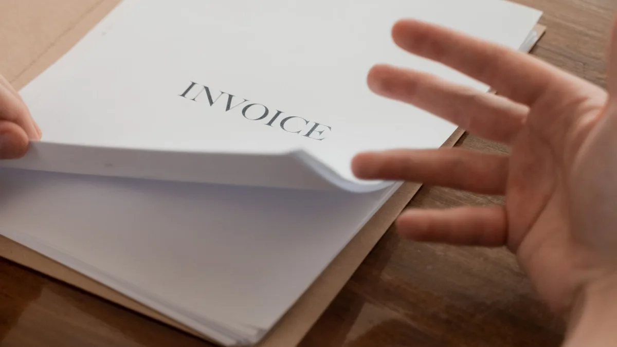 Una mano abre un documento en papel con el encabezado "Invoice" sobre una mesa de madera