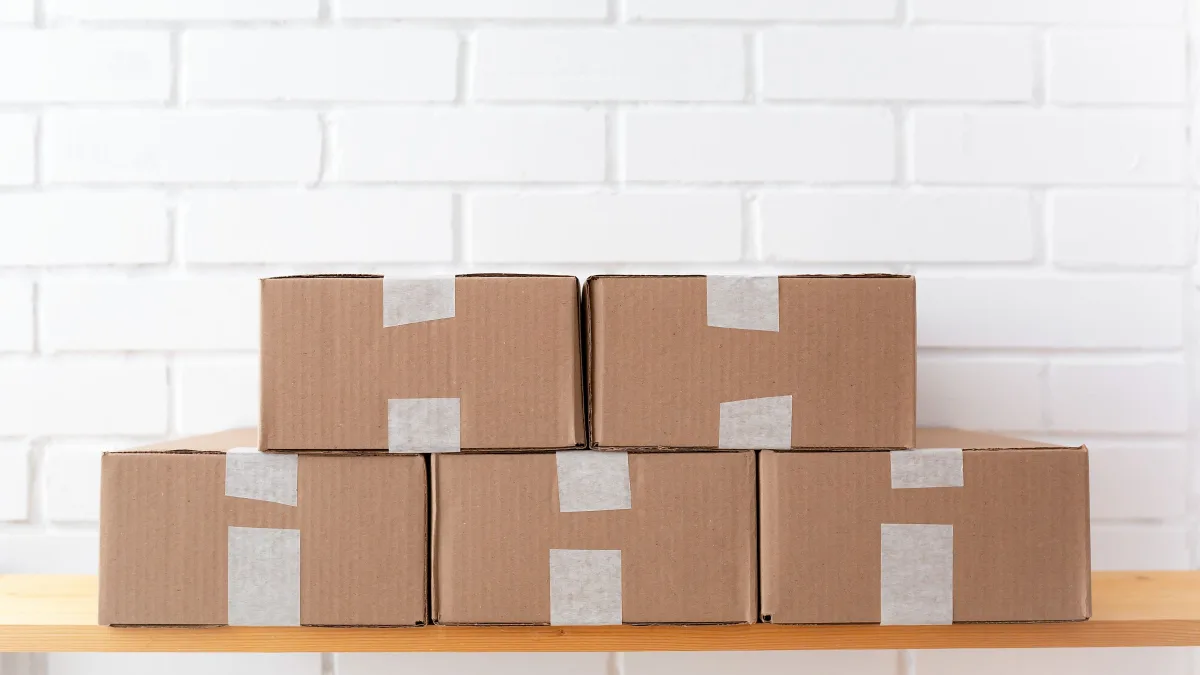Cajas de cartón sin etiquetar apiladas sobre una estantería, delante de una pared de ladrillo blanco